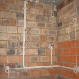 2Fire Piotr Matuszewski - Surowy mur z cegły dziurawki z rozprowadzonymi przewodami elektrycznymi w białych peszlach, widoczne puszki instalacyjne z wystającymi kablami w kolorze czerwonym i czarnym, brak wykończenia.