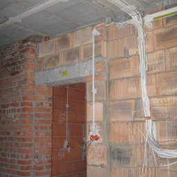 2Fire Piotr Matuszewski - Surowa instalacja elektryczna w trakcie montażu w nowym budynku z cegły, widoczne przewody prowadzone po ścianie i pod sufitem, puszki elektryczne w ścianie.