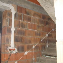 2Fire Piotr Matuszewski - Murowana ściana z cegły dziurawki w trakcie montażu instalacji elektrycznej: przewody prowadzone po powierzchni ściany, zabezpieczone gipsem, widoczne kable wychodzące z puszki elektrycznej.