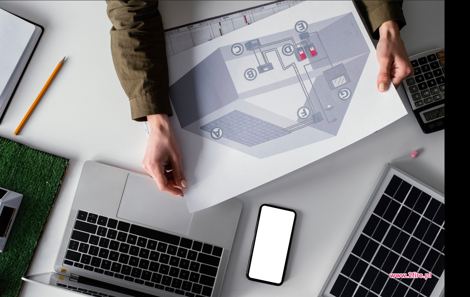 Osoba trzyma schemat instalacji fotowoltaicznej nad biurkiem z laptopem, kalkulatorem, panelem słonecznym i modelem domu z zielenią.