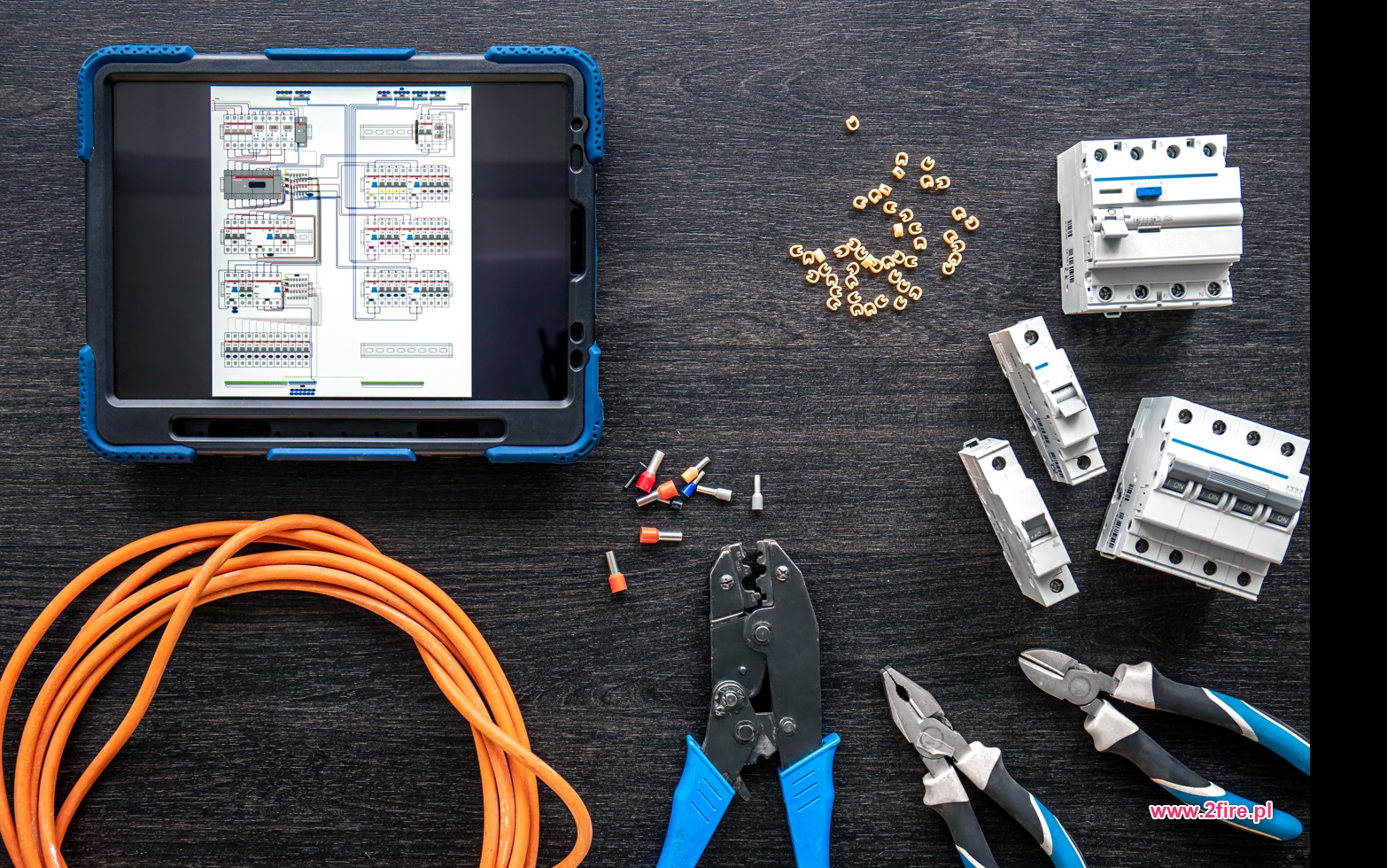 Tablet z schematem elektrycznym, przewód pomarańczowy, złączki, zaciskarka i wyłączniki na ciemnym drewnianym tle.