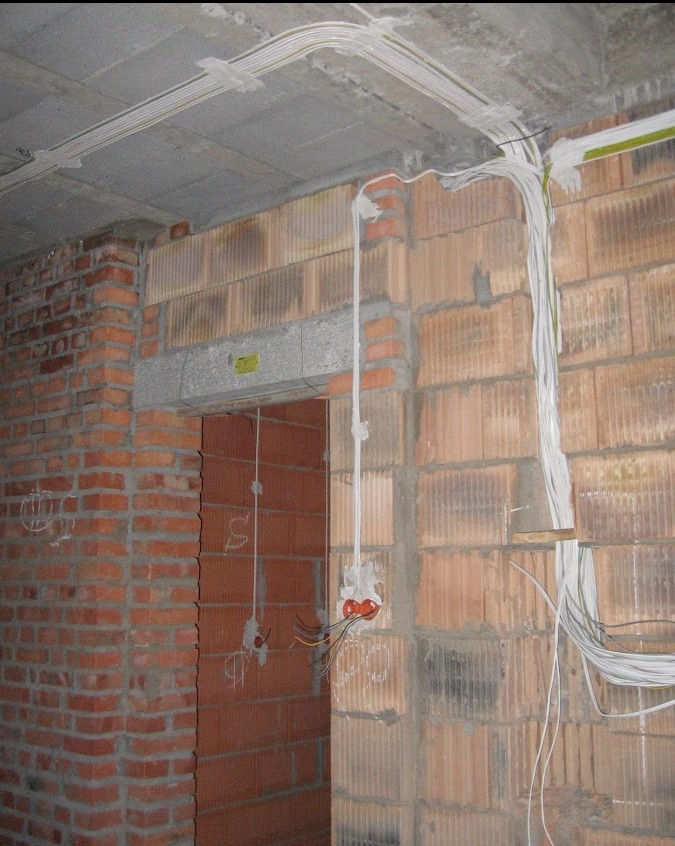 Surowa instalacja elektryczna w trakcie montażu w nowym budynku z cegły, widoczne przewody prowadzone po ścianie i pod sufitem, puszki elektryczne w ścianie.