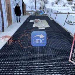 Przygotowanie podłoża pod kostkę brukową z użyciem plastikowej kratki drenażowej, widoczny pracownik w zimowej odzieży, częściowo ułożona kostka i narzędzia na placu budowy w zimowej scenerii.