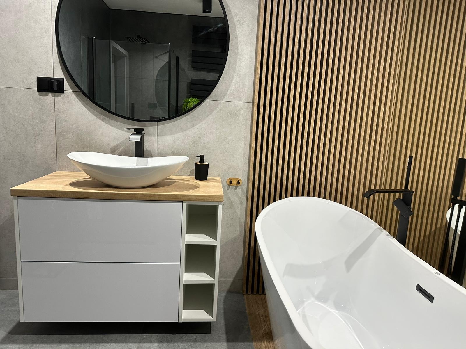 Nowoczesna łazienka z umywalką nablatową w kształcie misy na szafce z blatem drewnianym, okrągłe lustro z czarną ramą i drewniane lamele na ścianie w tle, obok wolnostojąca wanna.