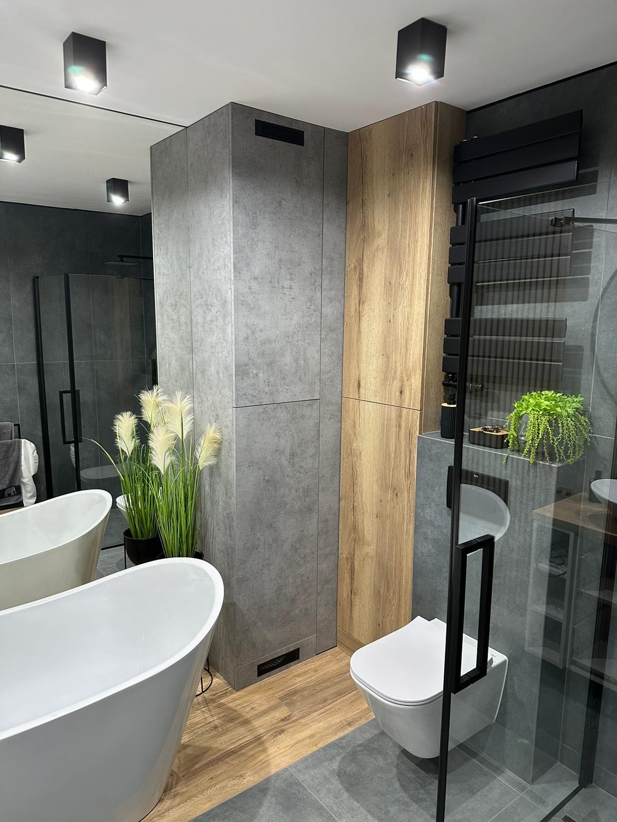 Nowoczesna łazienka z wolnostojącą białą wanną, szarą zabudową meblową imitującą beton, drewnianą szafką, czarną kabiną prysznicową i dekoracyjnymi roślinami w doniczkach.