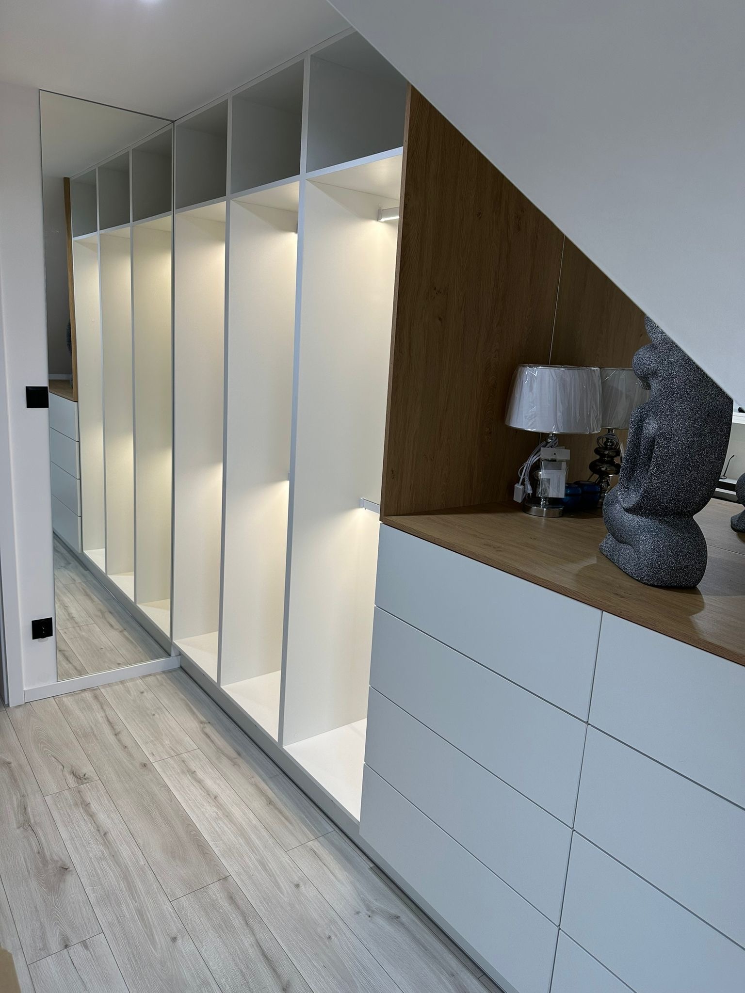 Biała szafa wnękowa z lustrzanymi drzwiami przesuwnymi i oświetleniem LED wewnątrz, dopasowana do skosu dachu, z drewnianym blatem, na którym stoją lampki nocne i dekoracyjna rzeźba.