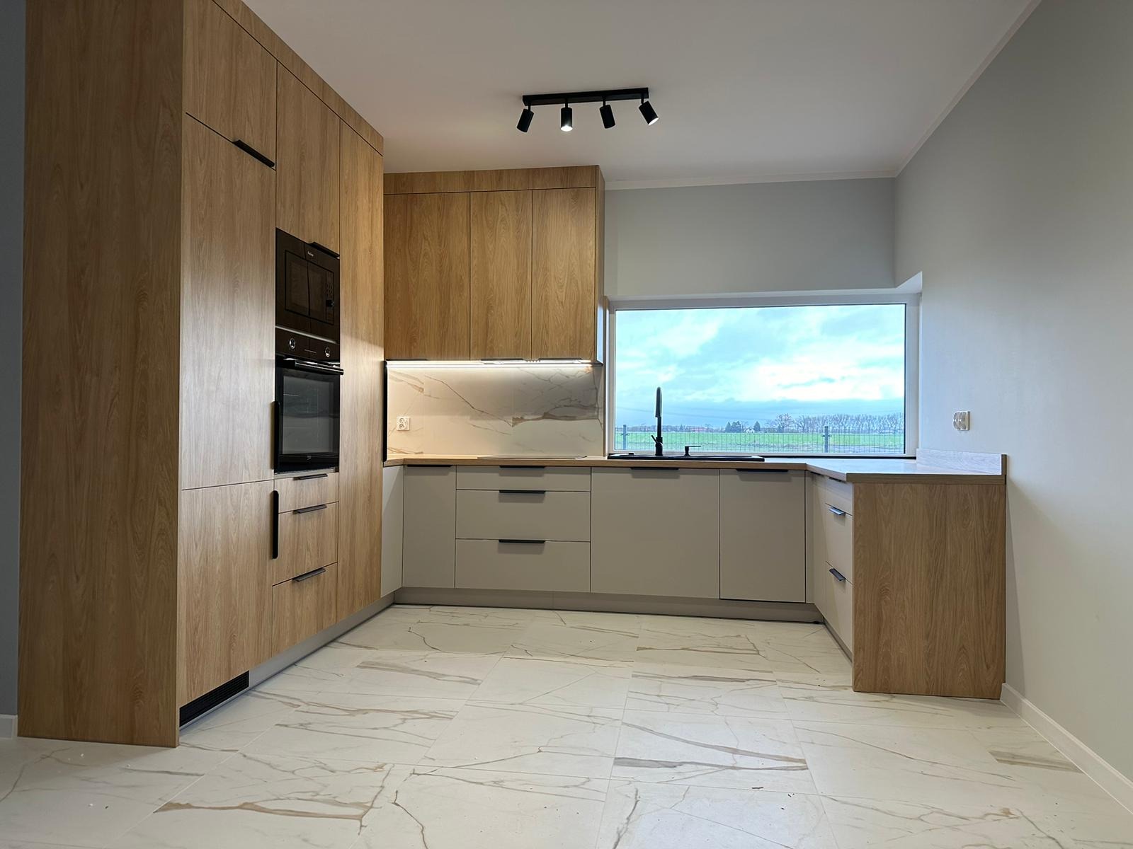 Jasna kuchnia z drewnianymi frontami szafek i szarymi dolnymi szafkami, marmurowa podłoga i widok na pole za oknem.