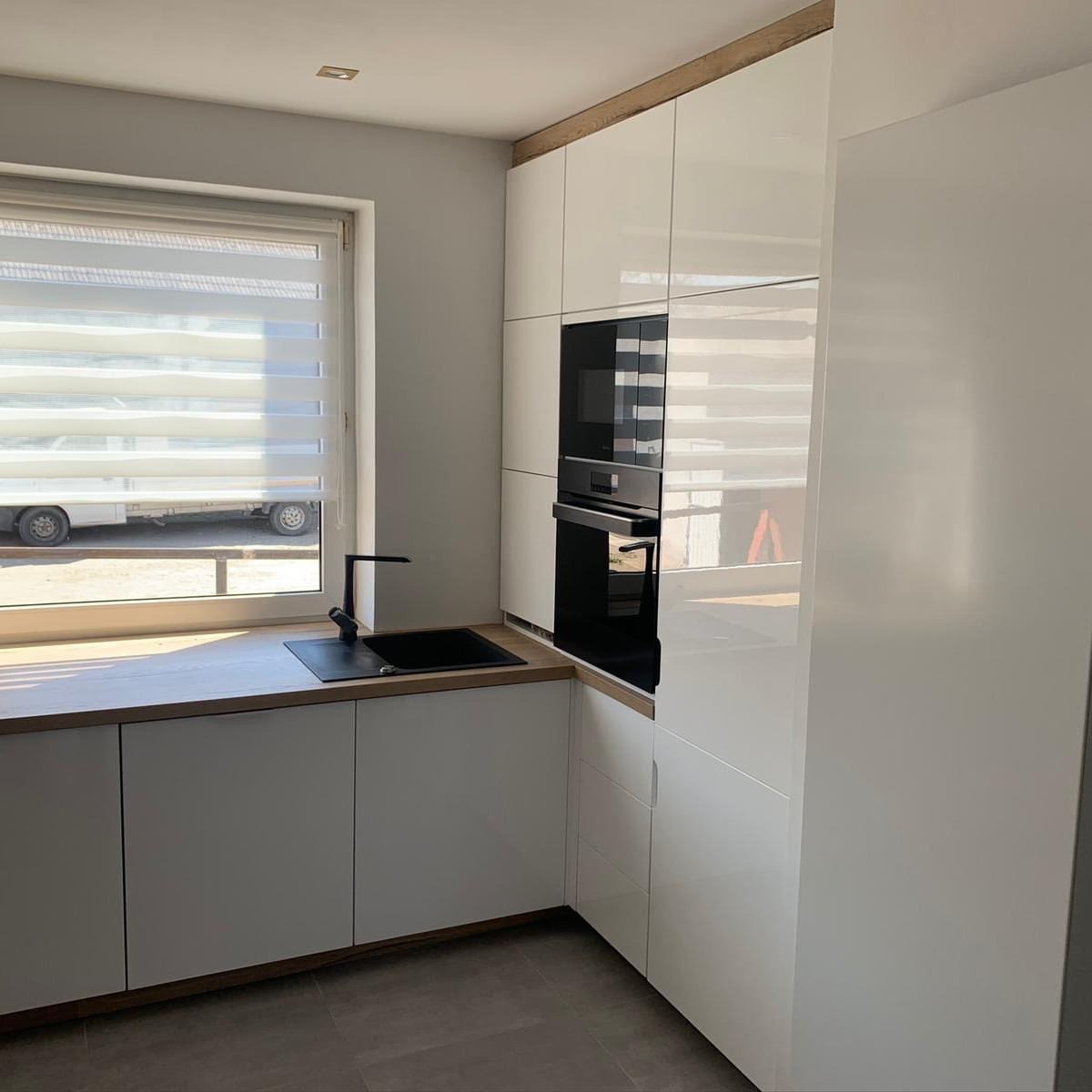 Nowoczesna kuchnia z białymi, lakierowanymi frontami szafek, drewnianym blatem i czarnym zlewem pod oknem z roletami poziomymi; widoczny również wbudowany piekarnik.