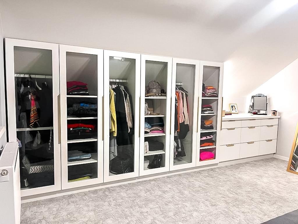 Biała szafa wnękowa z przeszklonymi drzwiami i złotymi uchwytami, zorganizowana odzież widoczna w środku, w pomieszczeniu na poddaszu w Święciechowie.