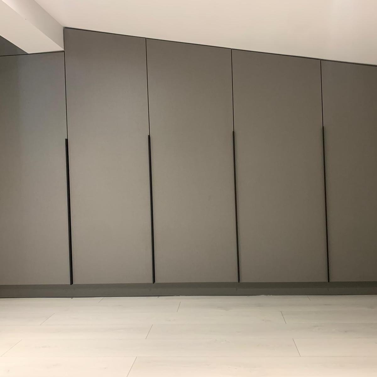 Zabudowa ściany szafami z szarymi frontami i minimalistycznymi, pionowymi, czarnymi uchwytami, na podłodze jasne panele.