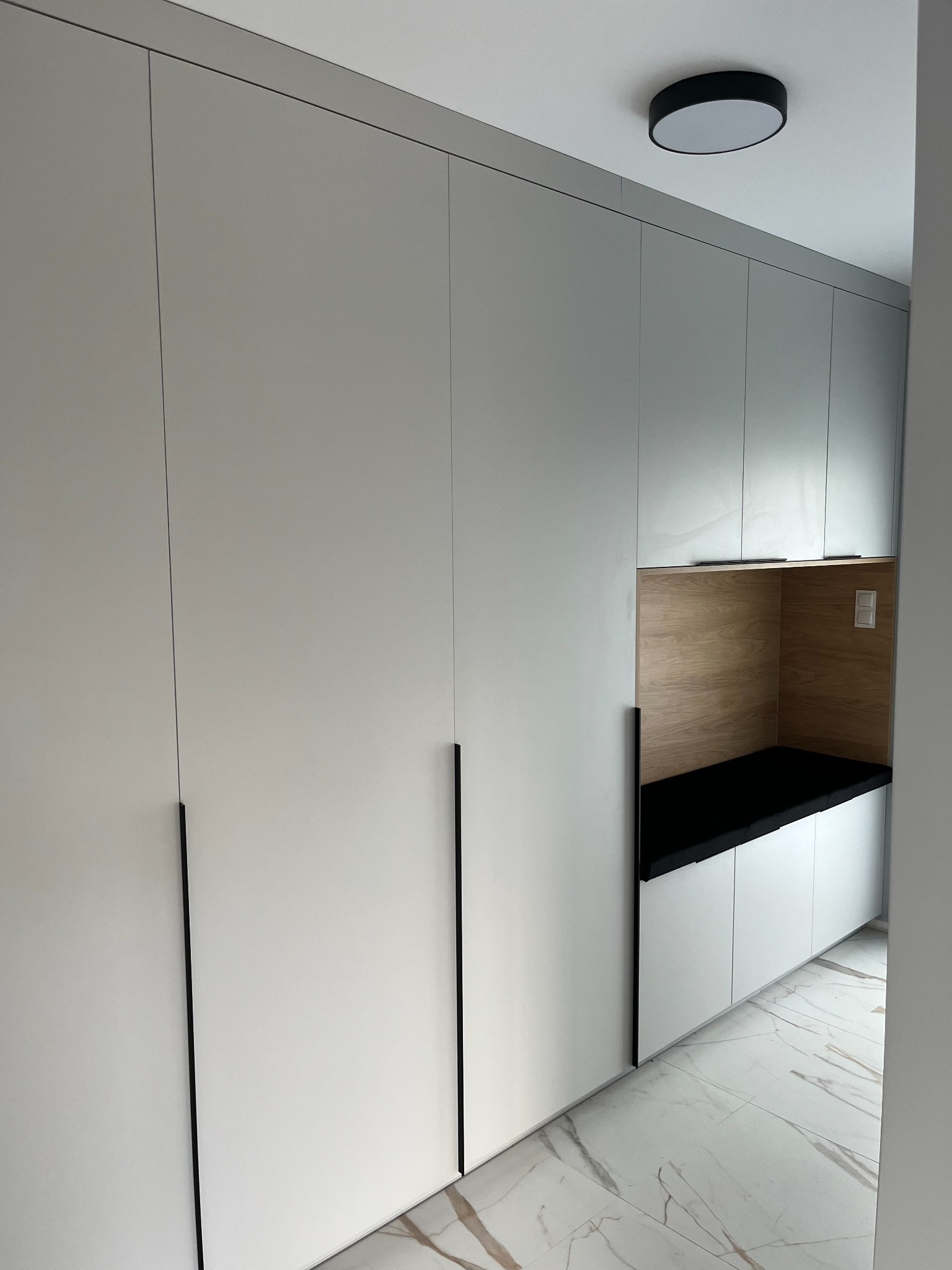 Biała szafa wnękowa z długimi, czarnymi uchwytami i wnęką z drewnianym wykończeniem oraz czarnym siedziskiem, oświetlona okrągłą lampą sufitową.