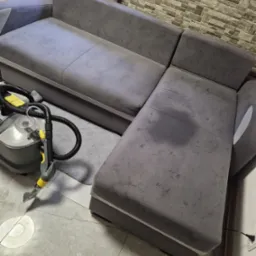 Szara narożna kanapa z widocznym mokrym śladem po praniu tapicerki, obok stoi odkurzacz piorący Karcher z żółtymi elementami, na podłodze marmurowe płytki.