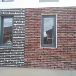 Fragment elewacji budynku z dwoma oknami, wykończony cegłą klinkierową w dwóch kolorach: czarnym i czerwonym. W oknach odbija się sąsiedni budynek i samochód.