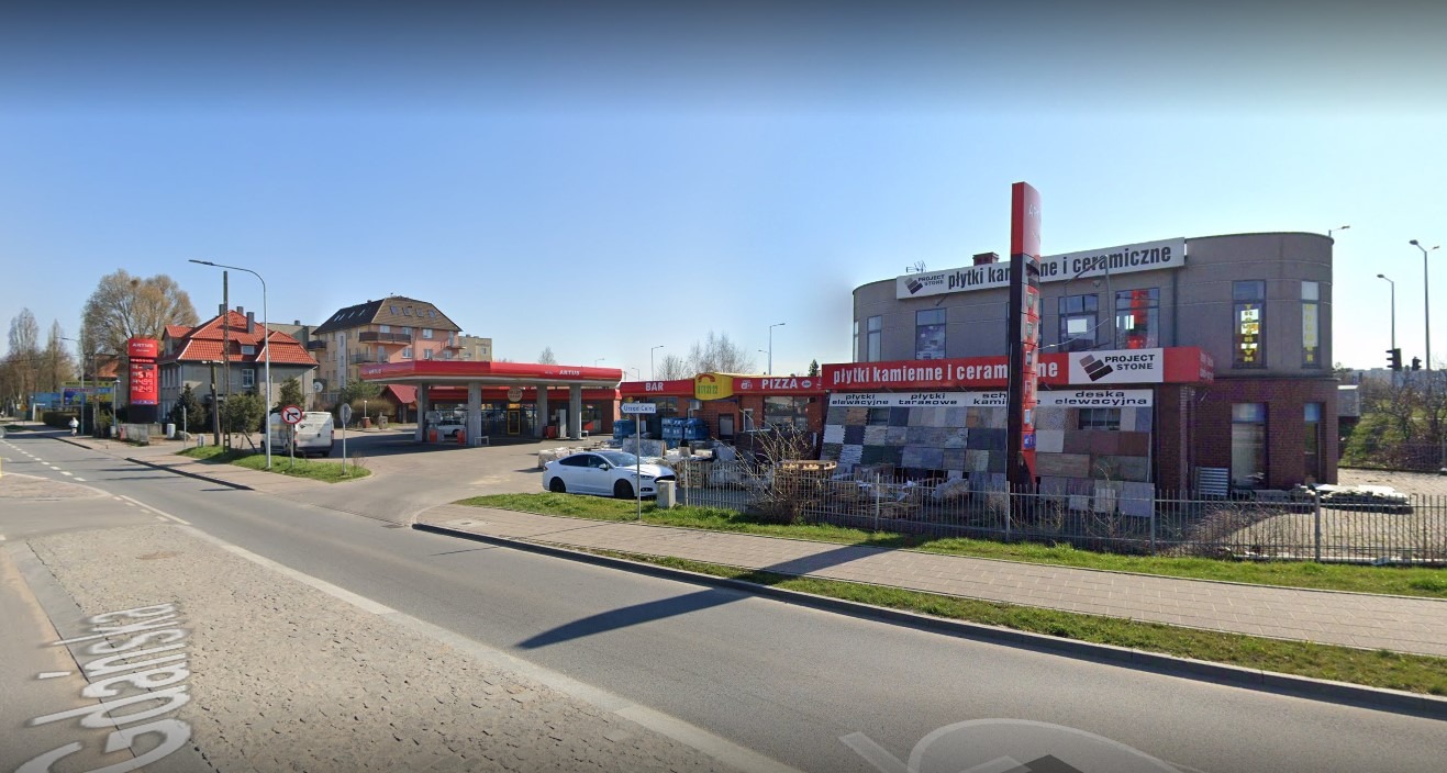 Widok na budynek firmy Project Stone z ekspozycją płytek kamiennych i ceramicznych na zewnątrz, widoczne reklamy i otoczenie miejskie.