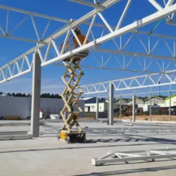 Montaż białej konstrukcji stalowej hali z użyciem podnośnika nożycowego na tle błękitnego nieba. Widoczny pracownik w żółtej kamizelce.