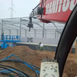 Montaż płyt warstwowych na konstrukcji stalowej hali z użyciem podnośnika pr&oacute;żniowego. Widok z kabiny operatora, w tle słup wysokiego napięcia.