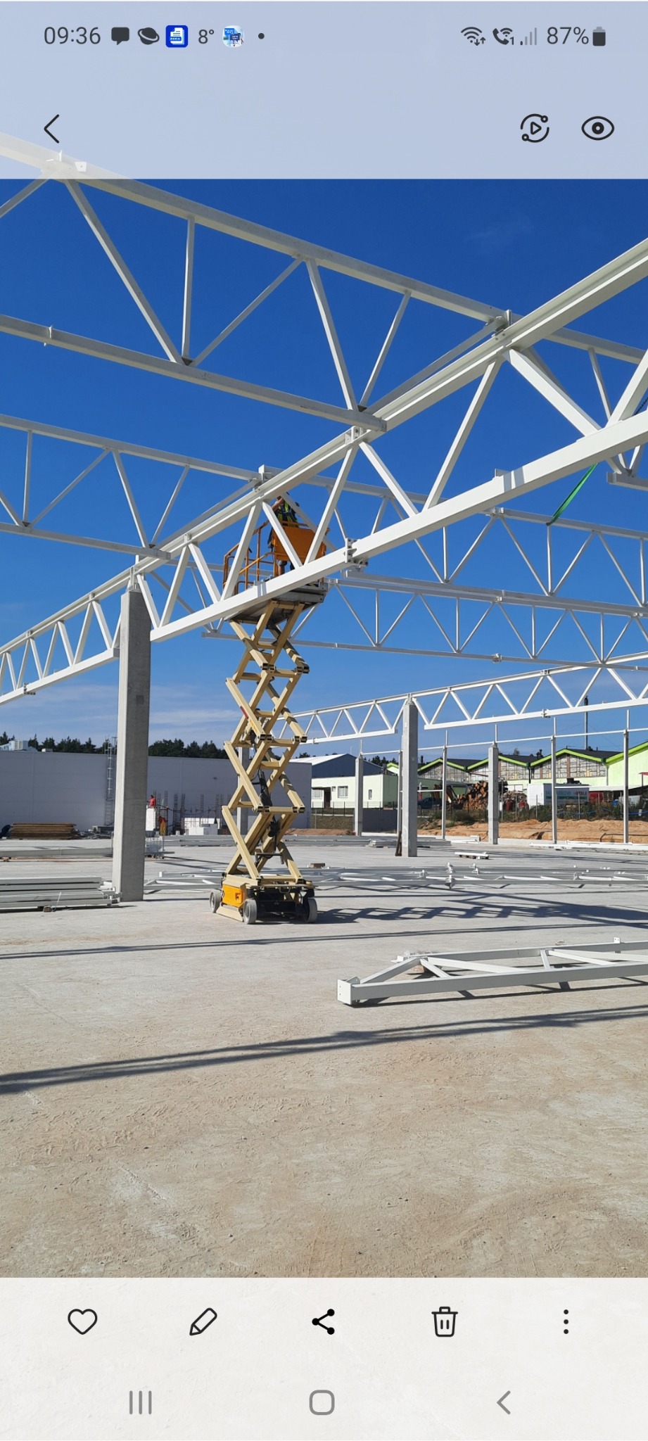 Montaż białej konstrukcji stalowej hali z użyciem podnośnika nożycowego na tle błękitnego nieba. Widoczny pracownik w żółtej kamizelce.