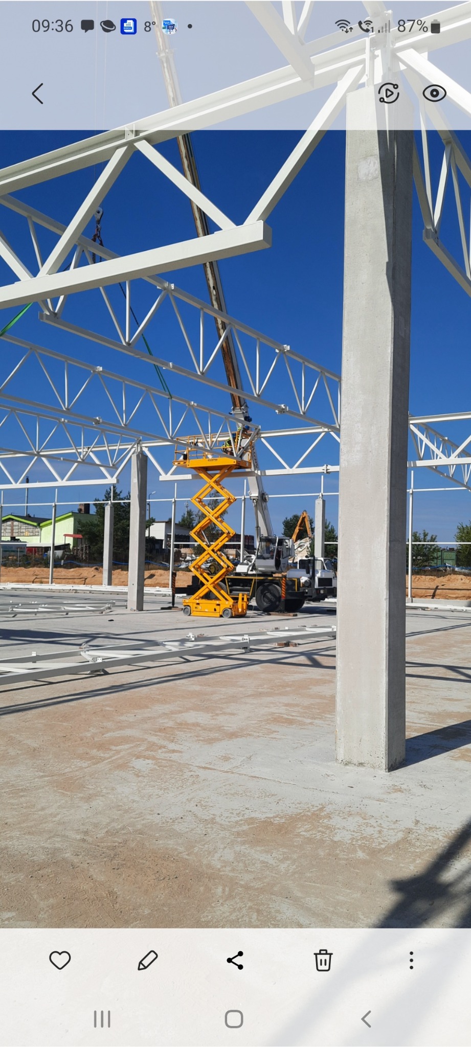 Montaż konstrukcji stalowej hali z użyciem podnośnika nożycowego, widoczne elementy konstrukcyjne i betonowe słupy nośne w słoneczny dzień.