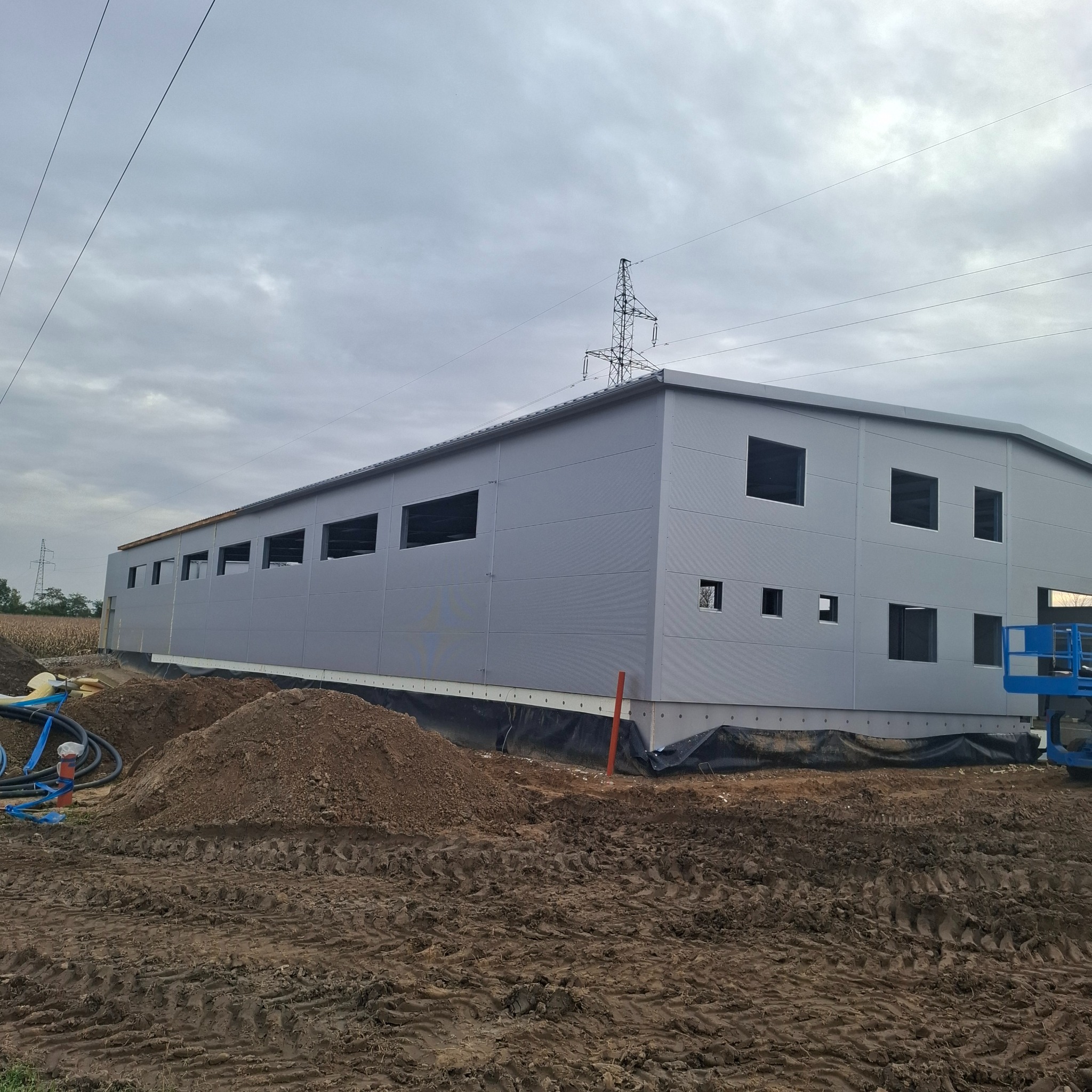 Budynek przemysłowy w trakcie montażu z płyt warstwowych, widoczny z zewnątrz, z przygotowanym terenem i elementami instalacji. Pochmurne niebo w tle.