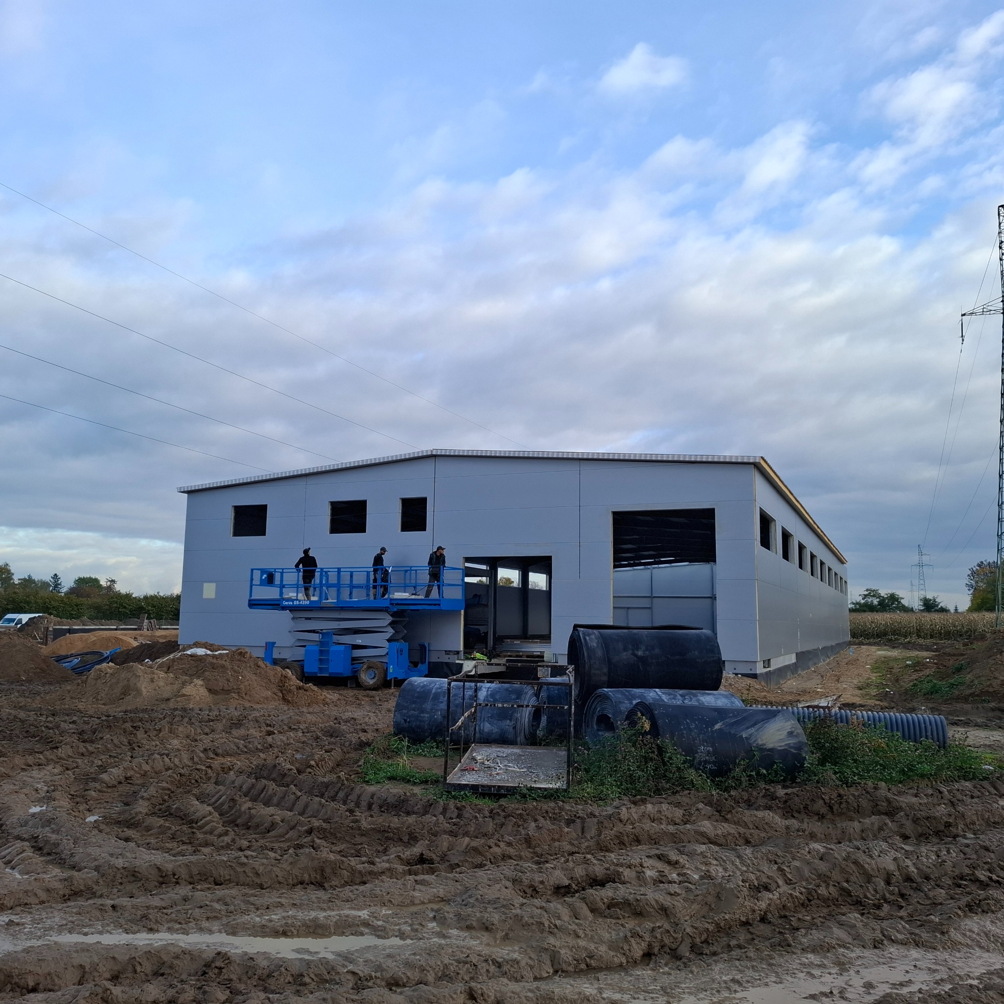 Montaż hali z płyt warstwowych: ekipa na podnośniku montuje elementy. Widoczne płyty, konstrukcja, teren budowy i niebo z chmurami. Prace w toku.