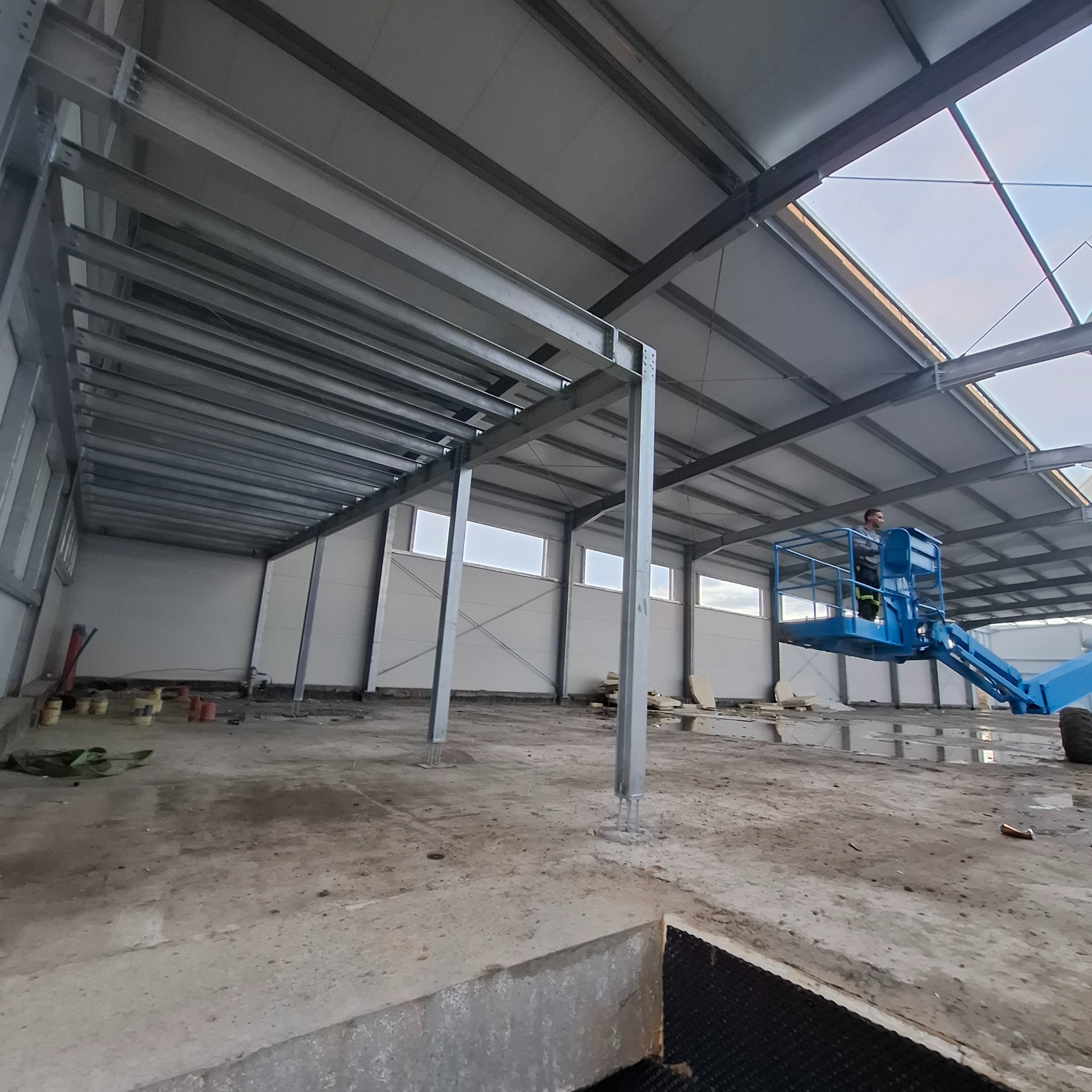 Wnętrze hali w trakcie montażu płyt warstwowych. Widoczna konstrukcja stalowa, pracownik na podnośniku i częściowo zamontowane ściany z płyt. Surowy beton na podłodze.