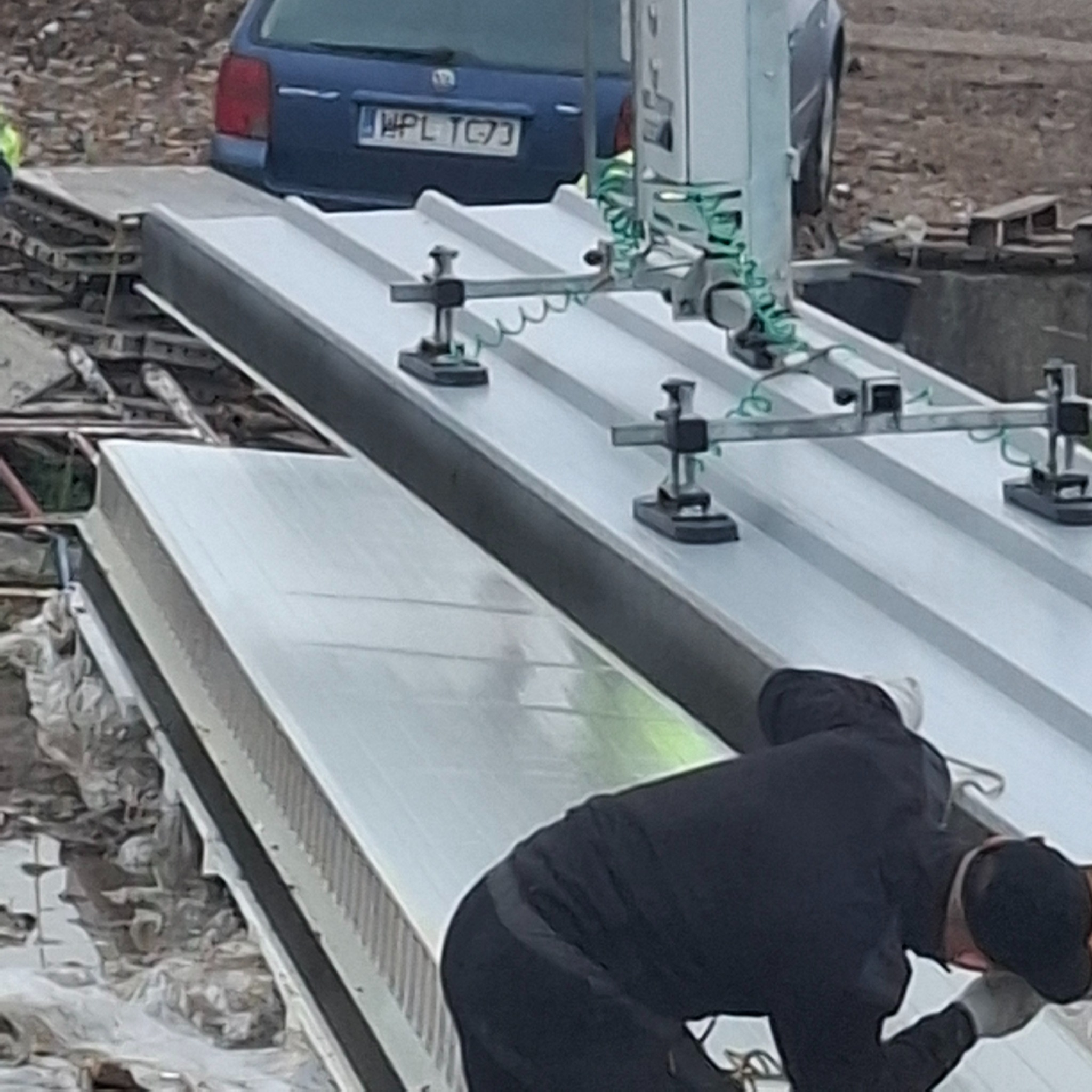 Montaż srebrnych płyt warstwowych za pomocą przyssawki. W tle widoczny fragment niebieskiego samochodu osobowego i elementy konstrukcji dachu.