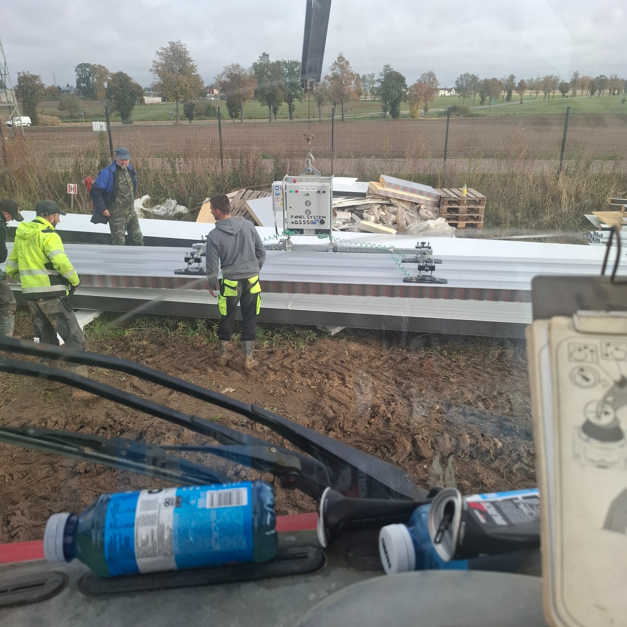 Montaż płyt warstwowych z użyciem przyssawki podnośnikowej. Widok z kabiny maszyny, w tle pracownicy i teren budowy.