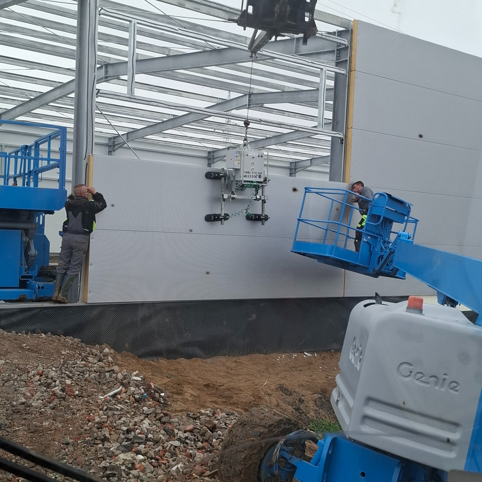 Montaż płyt warstwowych na konstrukcji stalowej przy użyciu podnośnika i specjalistycznego sprzętu. Dwóch pracowników nadzoruje proces.