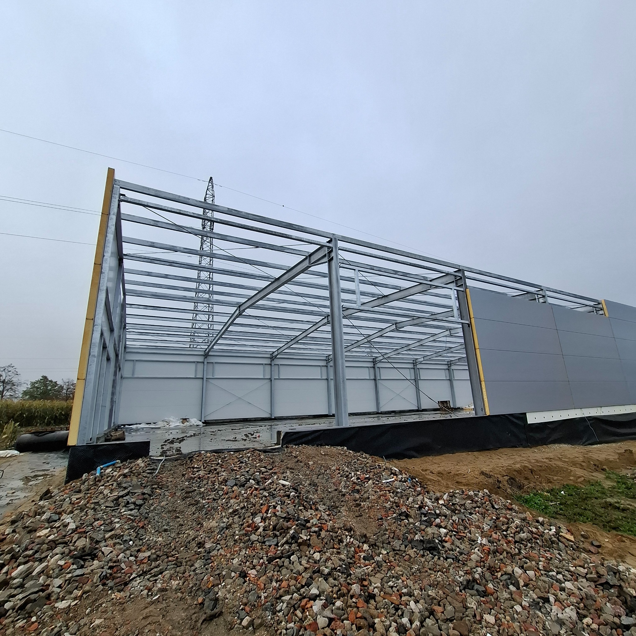 Konstrukcja stalowa hali w trakcie montażu płyt warstwowych, widoczne elementy szkieletu i fragmenty obudowy. Na pierwszym planie gruz i ziemia.