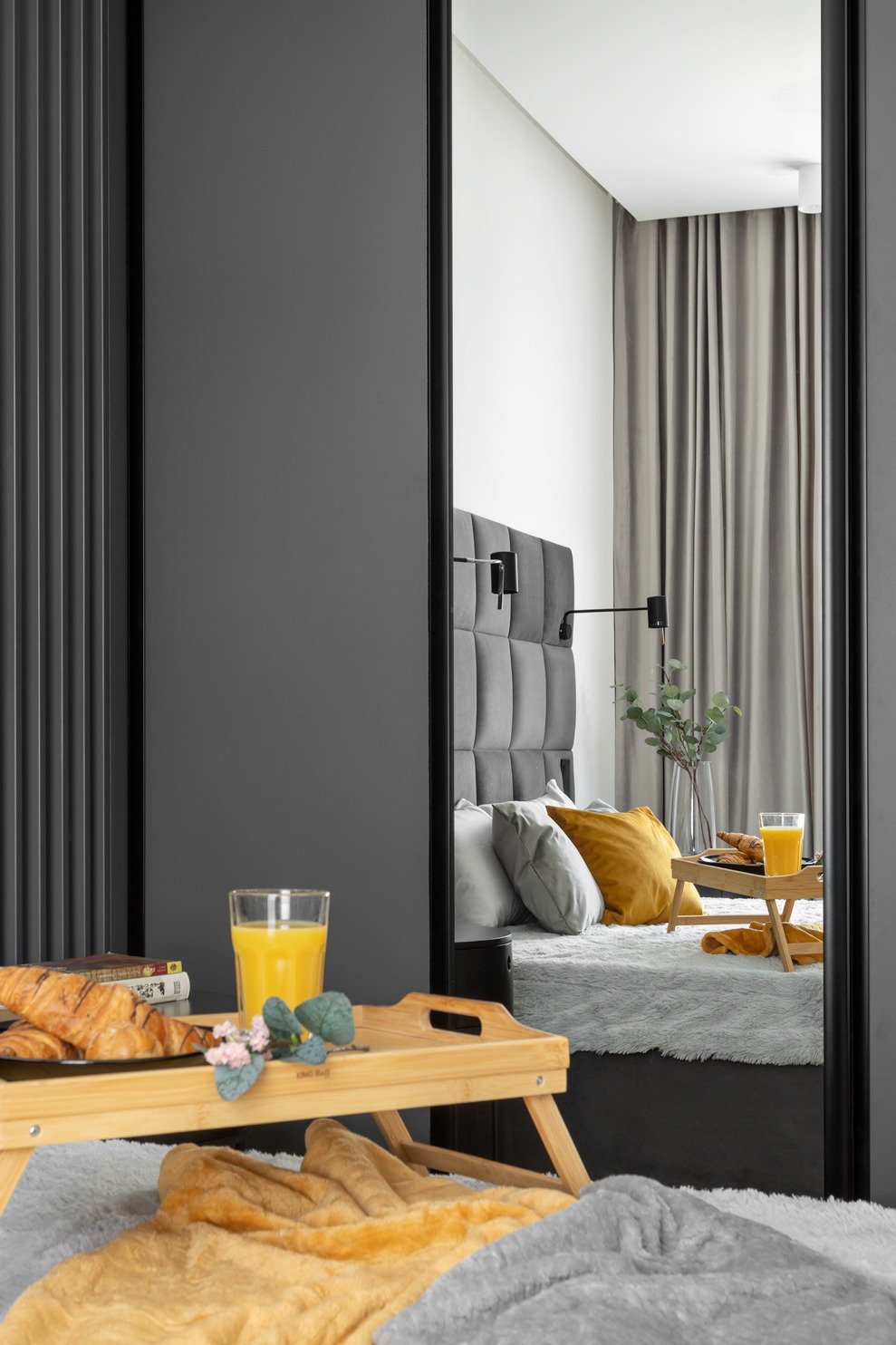 Sypialnia z śniadaniem na tacy, widoczna w lustrze z czarną ramą, z detalami wystroju w odcieniach szarości i żółci.