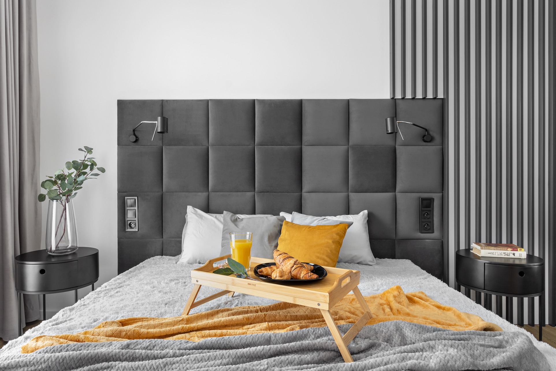 Nowoczesna sypialnia z szarym pikowanym zagłówkiem, drewnianym stolikiem śniadaniowym z croissantami i sokiem, na tle pionowych paneli dekoracyjnych.