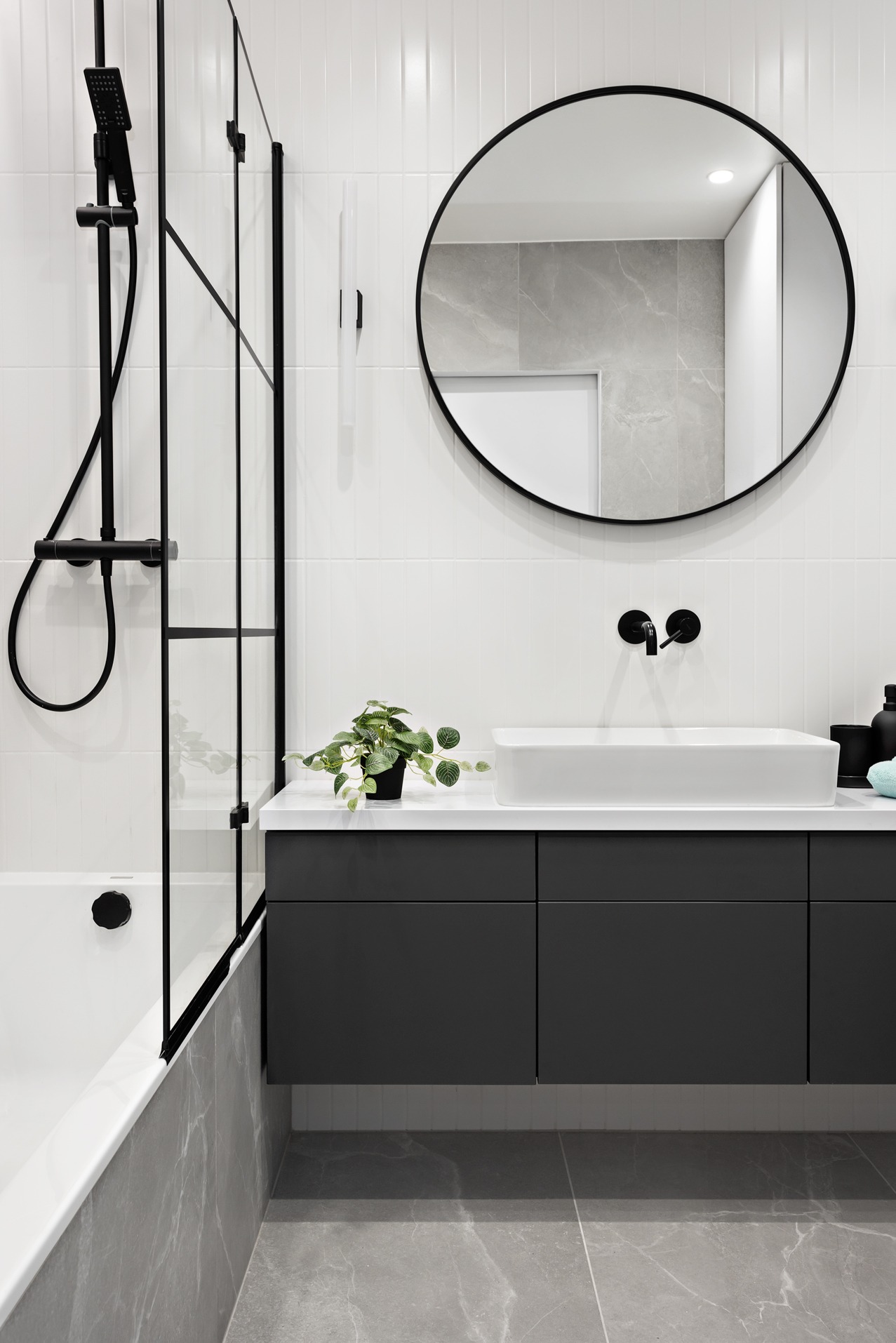 Nowoczesna łazienka z czarnymi akcentami: kabina prysznicowa typu walk-in, okrągłe lustro, umywalka nablatowa na szarej szafce i białe płytki na ścianach.