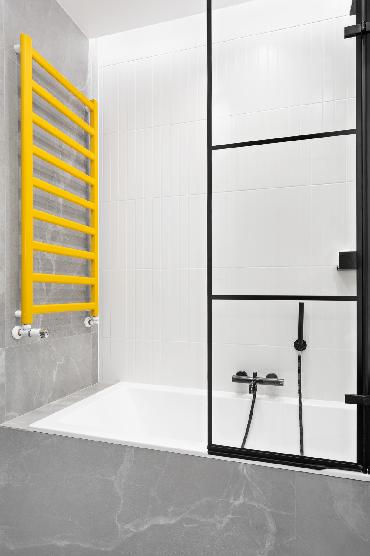 Nowoczesna łazienka z wanną obudowaną szarą płytką imitującą marmur, żółty grzejnik drabinkowy i czarna kabina prysznicowa z minimalistycznymi profilami.