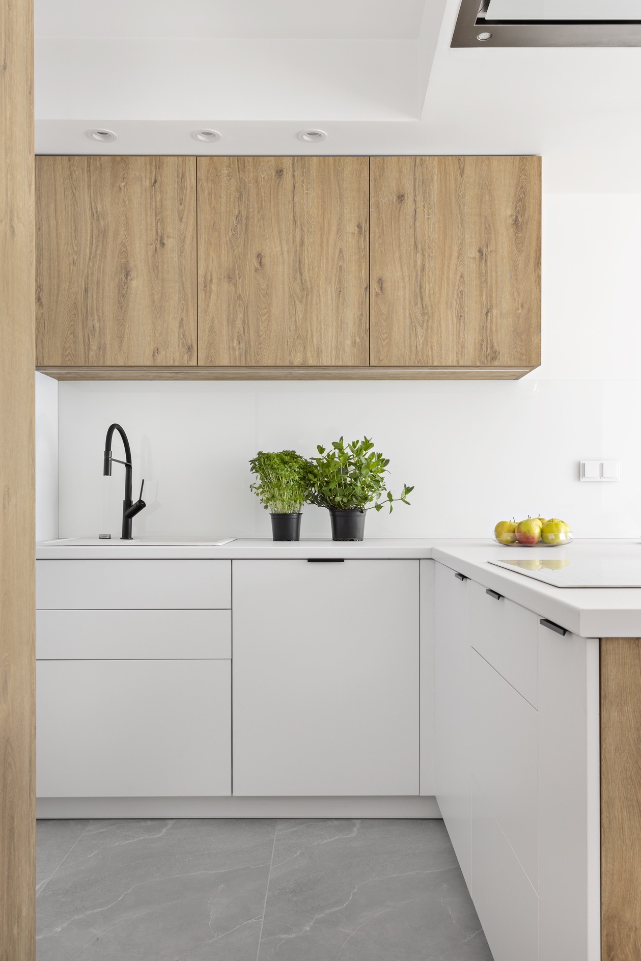 Nowoczesna kuchnia z białymi szafkami dolnymi i drewnopodobnymi górnymi, czarna bateria kuchenna, dwie doniczki z ziołami i jabłka na blacie.
