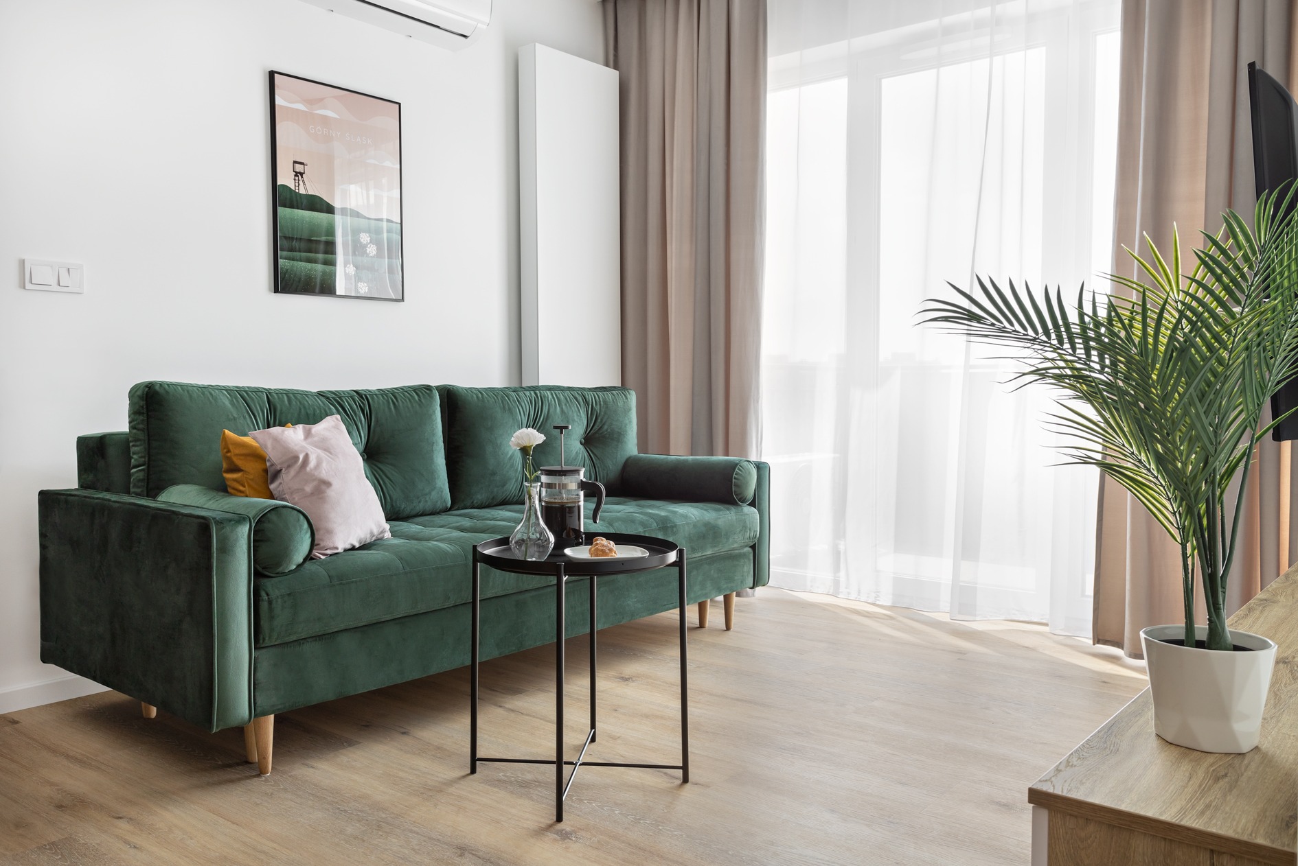Szmaragdowa sofa z aksamitnym obiciem, poduszki w odcieniach różu i musztardy, stolik kawowy z czarnym blatem i kwiatem w wazonie, jasna podłoga i naturalne światło z okna.