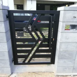 Nowoczesna, czarna brama z poziomymi przęsłami i geometrycznym wzorem, zamontowana między szarymi, betonowymi słupkami. W tle widoczny fragment samochodu i budynku.