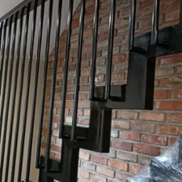 Nowoczesne schody metalowe z czarnymi, prostymi balustradami na tle ściany z czerwonej cegły, częściowo owinięte folią ochronną.