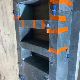 Home remont - Półka wnękowa wyłożona grafitową płytką, zabezpieczona pomarańczową taśmą i niebieskimi krzyżykami dystansowymi, widok na ścianę z panelami podłogowymi.