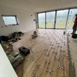 Home remont - Układanie płytek imitujących drewno w dużym pokoju z oknami, widoczne krzyżyki dystansowe, worki z zaprawą Axton, odkurzacz przemysłowy i pracownik w tle.