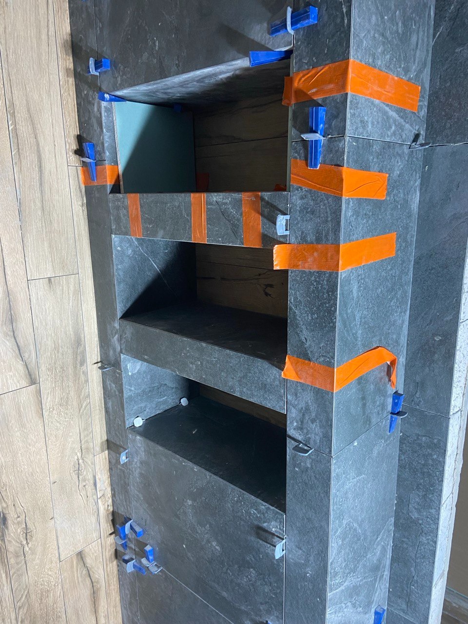 Półka wnękowa wyłożona grafitową płytką, zabezpieczona pomarańczową taśmą i niebieskimi krzyżykami dystansowymi, widok na ścianę z panelami podłogowymi.