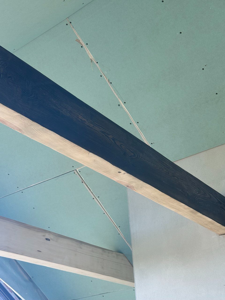 Montaż płyt gipsowo-kartonowych na suficie z widocznymi drewnianymi belkami konstrukcyjnymi, jedna z belek pomalowana na ciemny kolor.