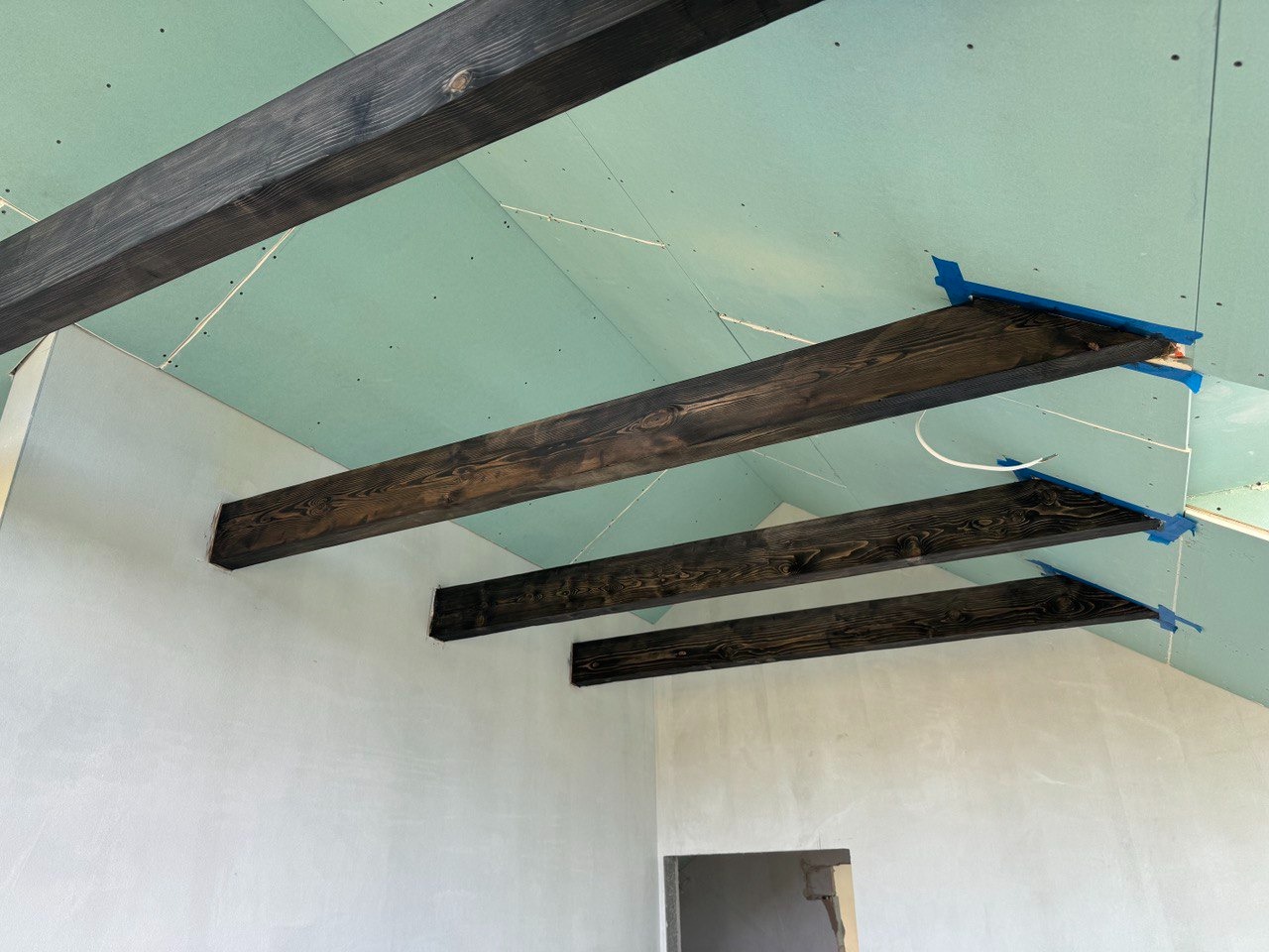 Montaż belek stropowych z ciemnego drewna pod skośnym sufitem z płyt gipsowo-kartonowych, zabezpieczonych niebieską taśmą malarską.