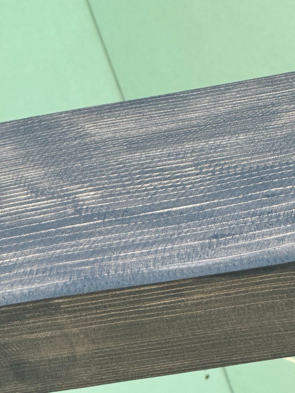 Drewniana listwa przypodłogowa w odcieniach szarości i błękitu, widoczne słoje drewna, na tle zielonej ściany.