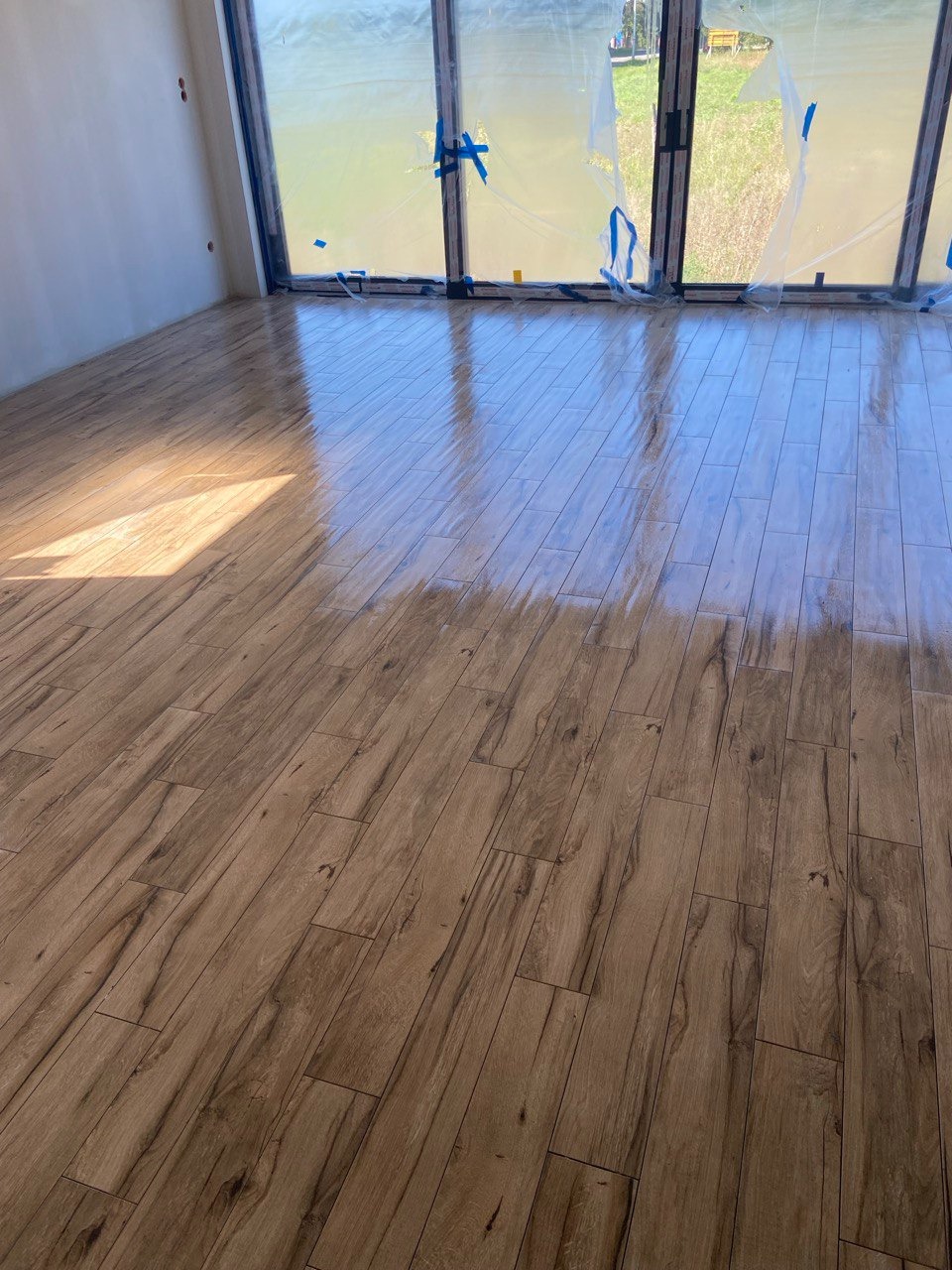 Nowo ułożona, lśniąca podłoga z paneli imitujących drewno w jasnym pomieszczeniu z dużym oknem zabezpieczonym folią malarską.