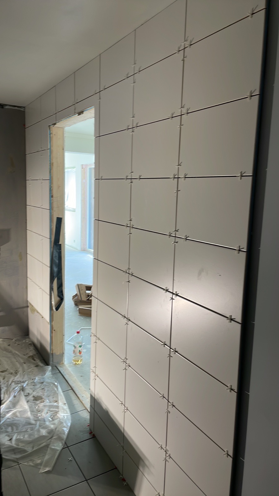 Ściana w trakcie układania białych, kwadratowych płytek, z krzyżykami dystansowymi, widoczny fragment ościeżnicy drzwiowej i folia ochronna na podłodze.