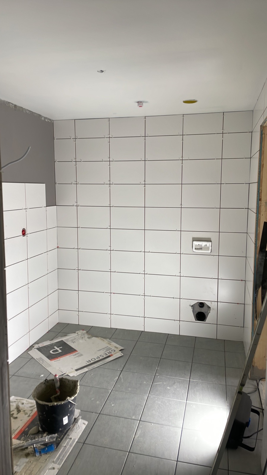 Łazienka w trakcie remontu: białe płytki ścienne z ciemną fugą, szare płytki podłogowe, wiadro z zaprawą, kartony po płytkach Grespor i widoczne instalacje.