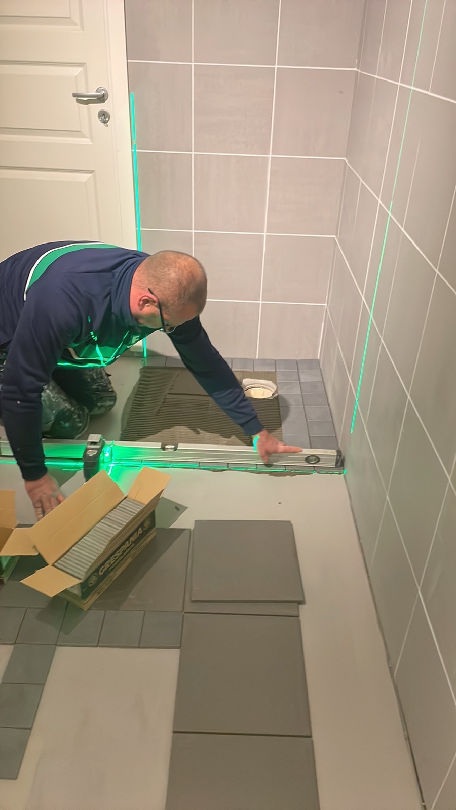 Układanie szarych płytek podłogowych w łazience z użyciem poziomicy laserowej; widoczna kartonowa paczka płytek Grespania i częściowo ułożona podłoga.