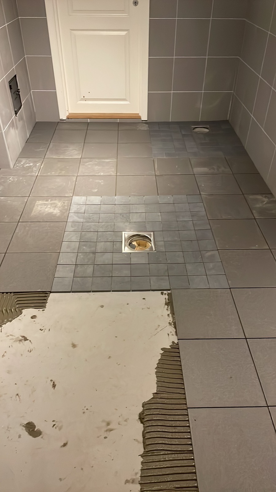 Szare płytki podłogowe w łazience, częściowo ułożone, z widoczną warstwą kleju i odpływem liniowym.