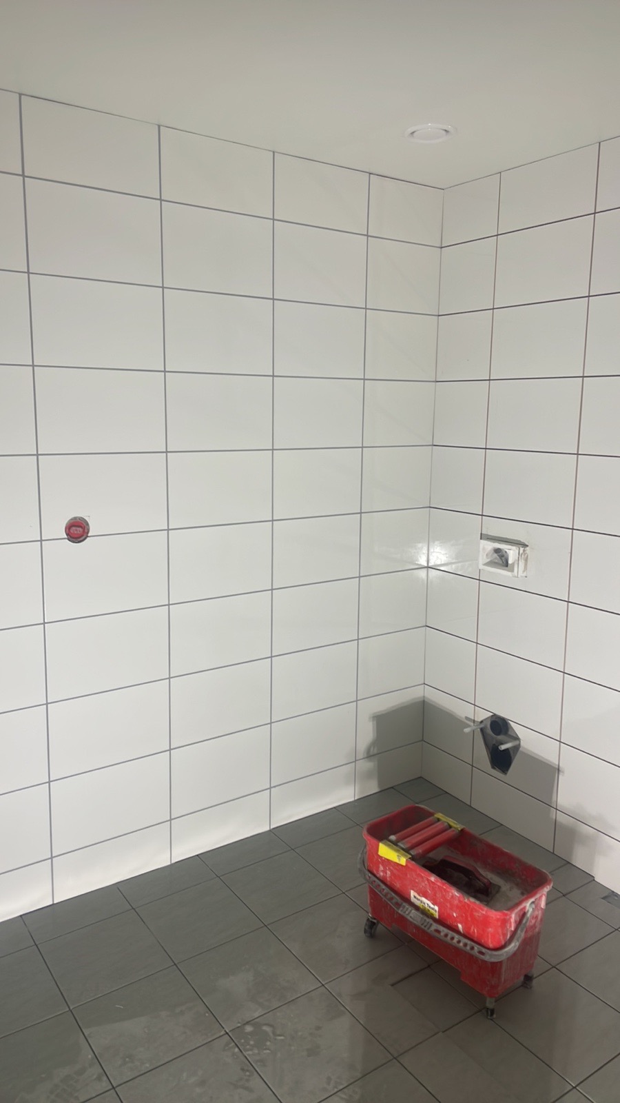Świeżo wykafelkowana łazienka z białą glazurą na ścianach i szarą terakotą na podłodze. Widoczny czerwony wózek z narzędziami do fugowania i resztki wody na podłodze.