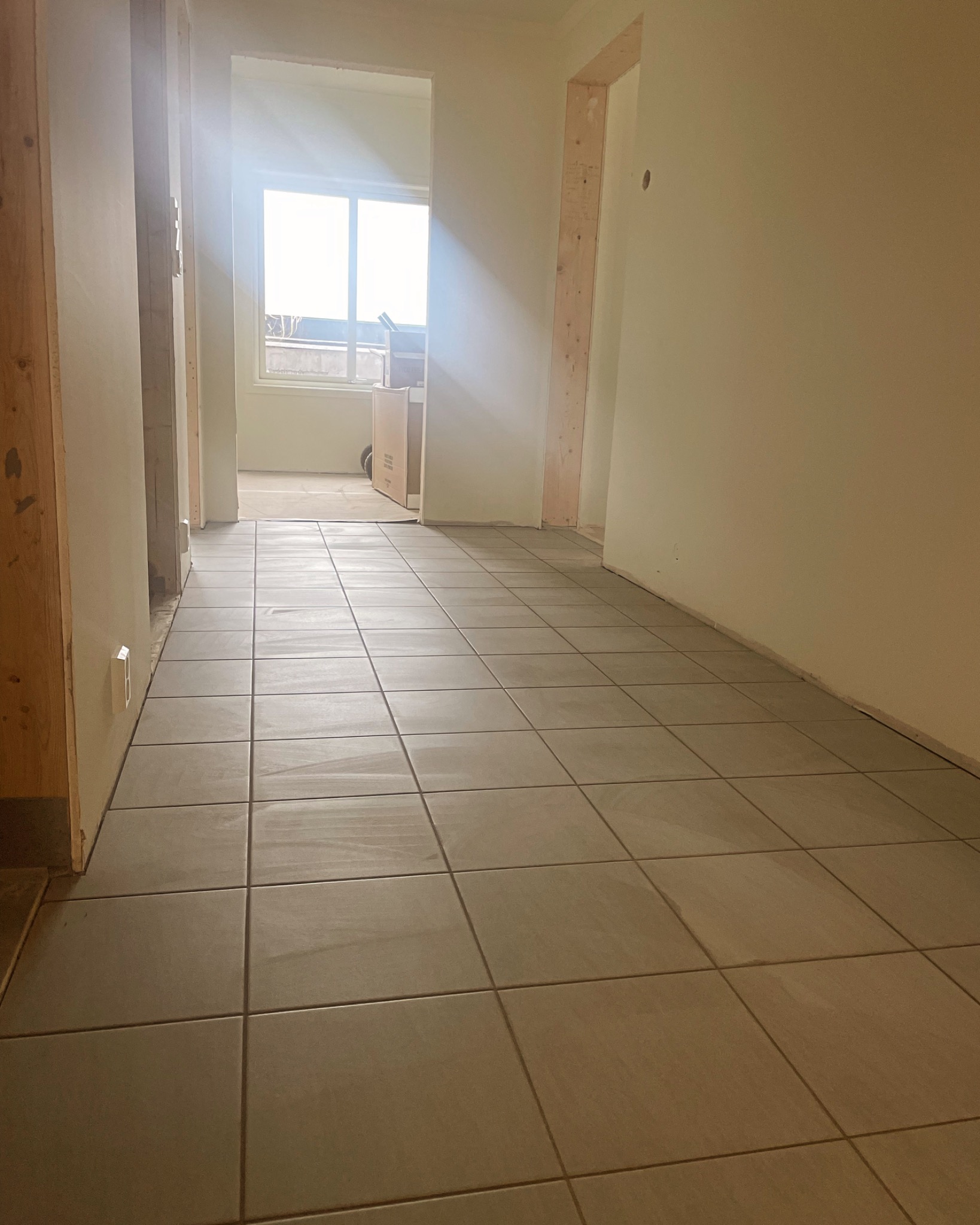 Korytarz w trakcie remontu z ułożonymi szarymi płytkami podłogowymi, surowe drewniane ramy drzwiowe, ściany przygotowane do malowania, widok na okno z kartonami w tle.